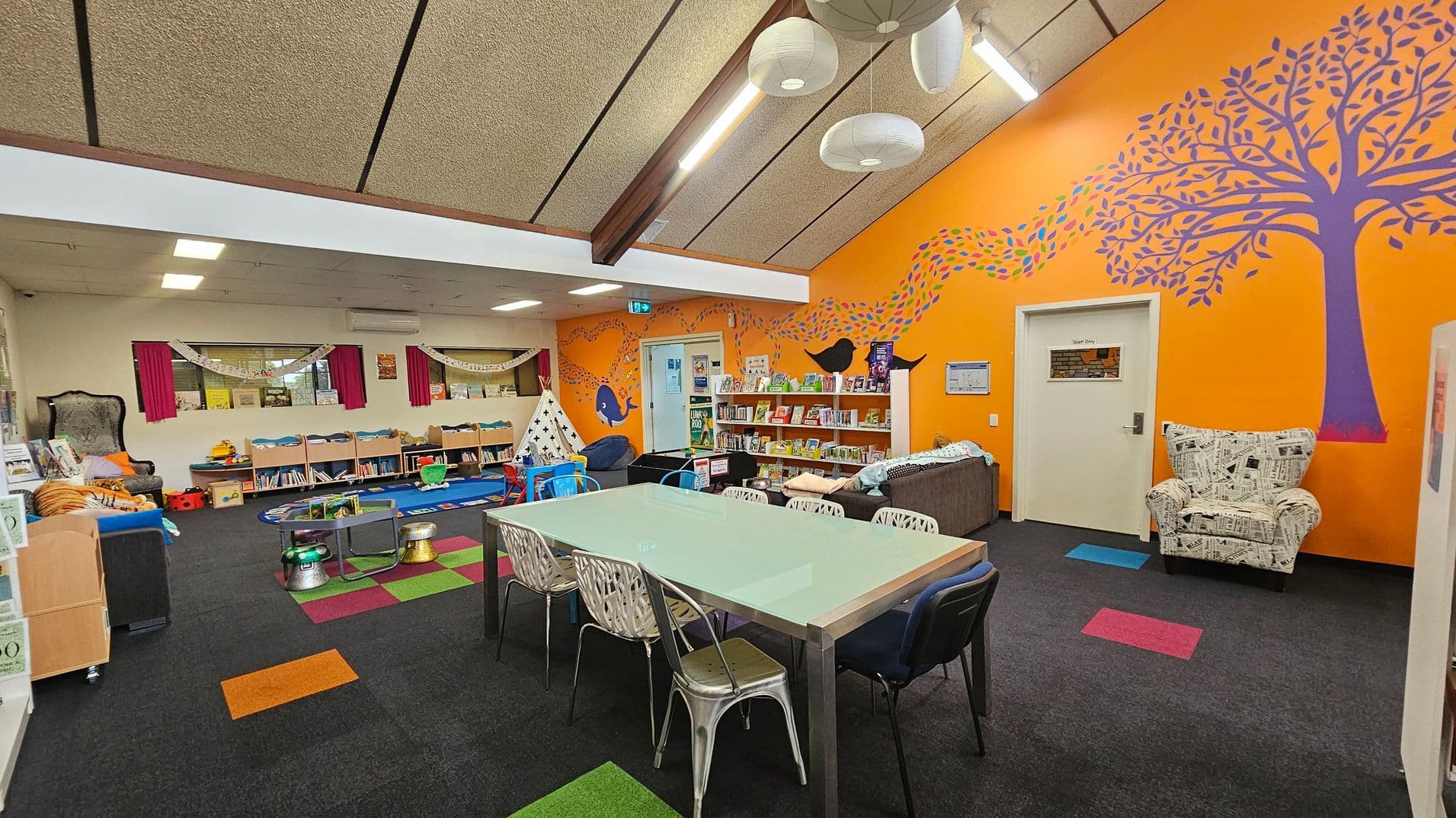 A colourful room with an orange wall on the right featuring a purple tree and whale mural. A white door and a bookshelf filled with books are also on this wall. In the centre, a large table with mismatched chairs sits on a dark carpet with colourful square patterns. On the left, a play area with a teepee, toys, and bookshelves is visible. The ceiling is sloped with exposed beams and hanging lights. The floor is carpeted in dark grey with scattered coloured squares.