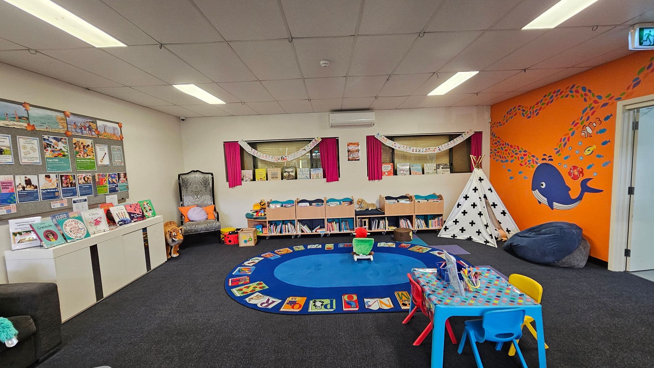 A colourful children's playroom with a dark carpeted floor. On the left, a white cabinet displays books and a bulletin board with posters above. A plush tiger toy sits beside a decorative chair. The centre features a blue circular rug with alphabet designs, surrounded by low bookshelves. On the right, an orange wall has a mural of a whale and fish. A white teepee and bean bag are nearby. A small table with colourful chairs is in the foreground. The ceiling has fluorescent lights.