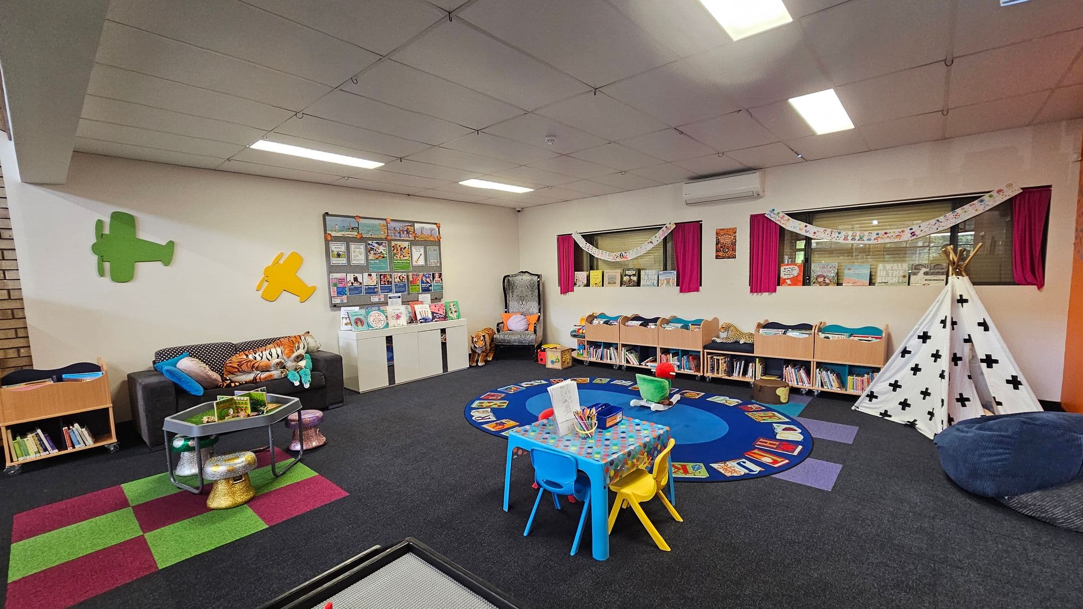 A colourful children's playroom with a black carpet. On the left, a grey sofa with plush toys and a small table with glittery stools. Green and yellow aeroplane wall decorations are above. In the centre, a blue circular rug surrounded by books. A small table with blue and yellow chairs is nearby. On the right, a white teepee with black crosses and a bean bag. Bookshelves line the wall, and a window with pink curtains is above. The ceiling has fluorescent lights.