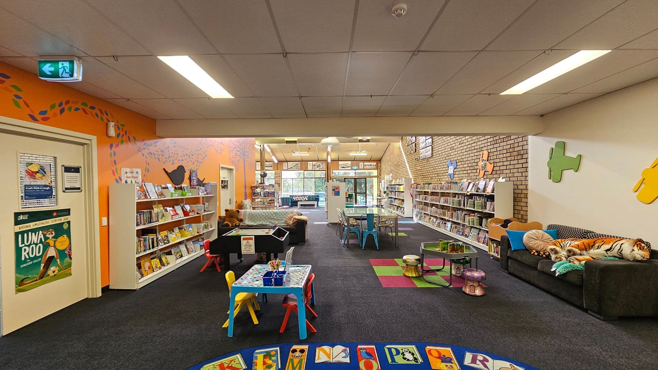 A colourful children's library area with an orange wall on the left featuring bookshelves filled with books and a poster. A small table with colourful chairs is in the centre, surrounded by a black carpet with an alphabet-themed rug. On the right, a dark sofa with plush toys, including a tiger, sits against a brick wall adorned with colourful plane cutouts. The ceiling has fluorescent lights, and the floor is carpeted with some green and red square patterns.
