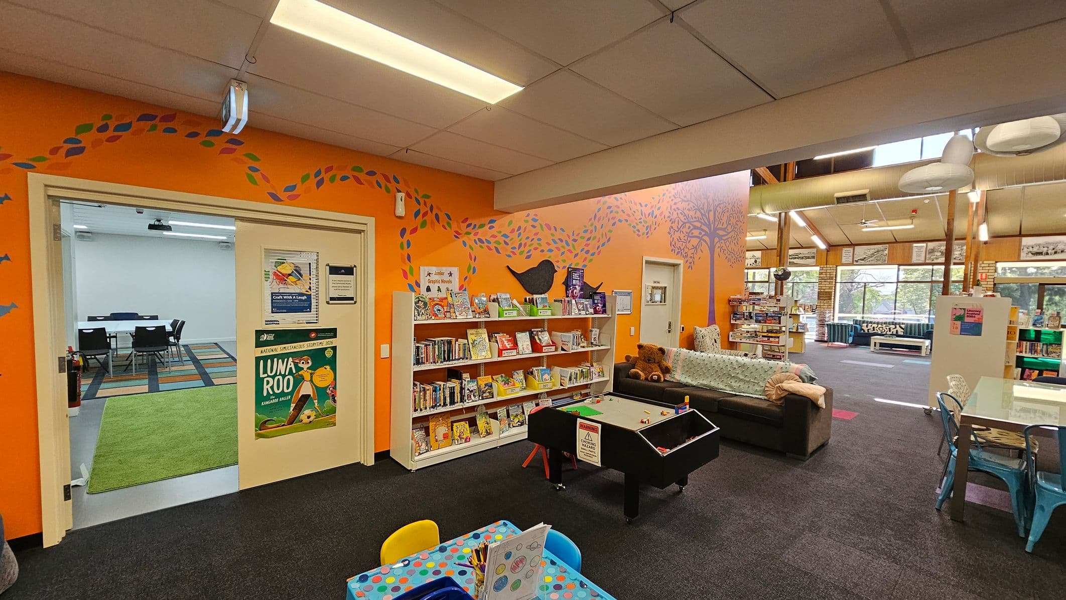 A library interior with an orange wall featuring a painted tree and bird design. On the left, there's a doorway leading to a room with a table and chairs on a striped carpet. Below the wall art, a bookshelf is filled with children's books. In the centre, a small game table is placed on a dark carpet. To the right, a sofa with cushions and a large teddy bear is visible. The background shows large windows and more seating areas with tables and chairs. The ceiling has fluorescent lights.