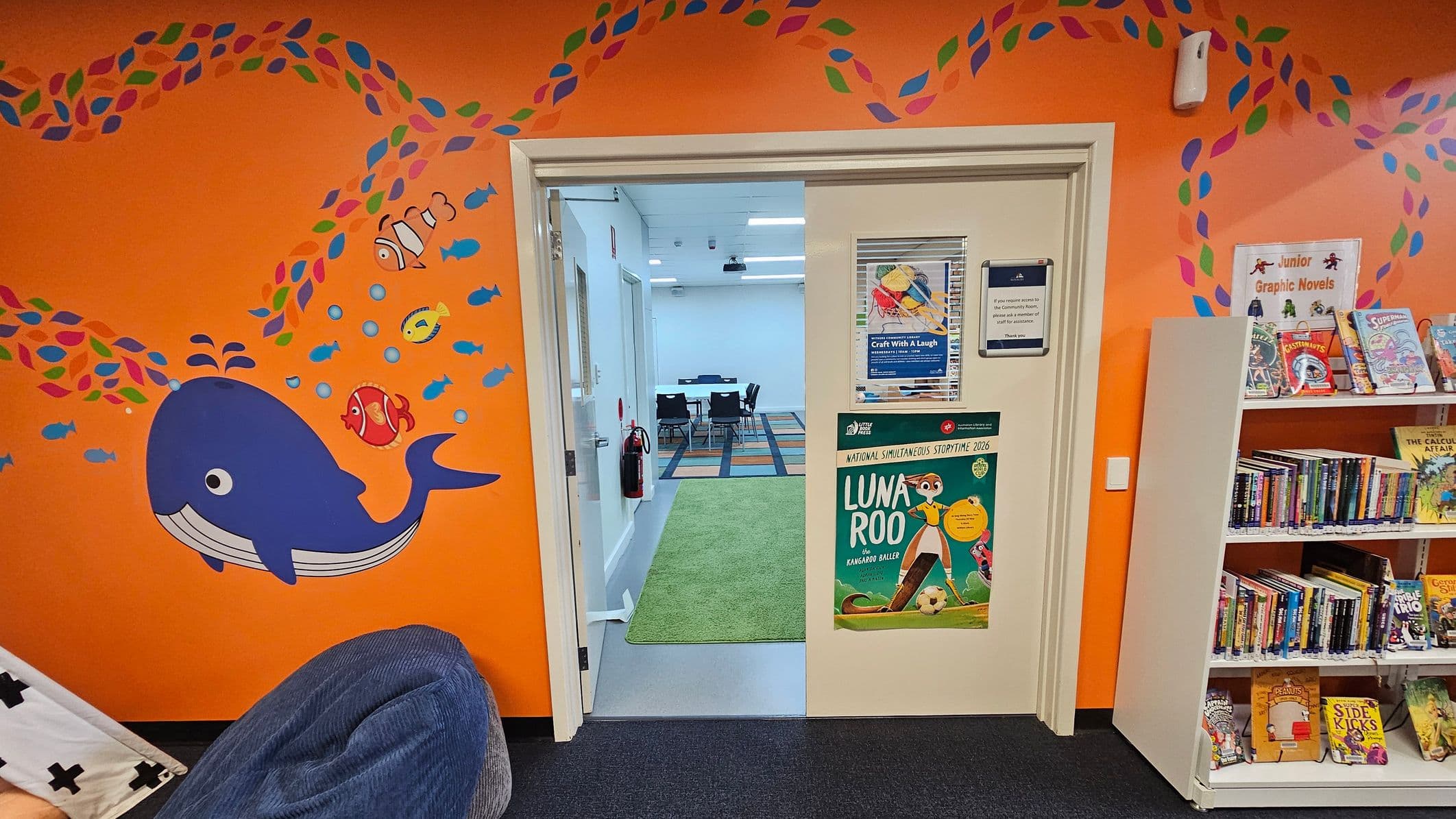 An orange wall with a colourful mural of a blue whale and fish on the left. In the centre, a doorway leads to a room with a green carpet and a table with chairs. Posters are displayed on the door, including one for "Luna Roo". On the right, a white bookshelf holds various children's graphic novels. The floor is carpeted in dark blue, and the ceiling is white with lights.