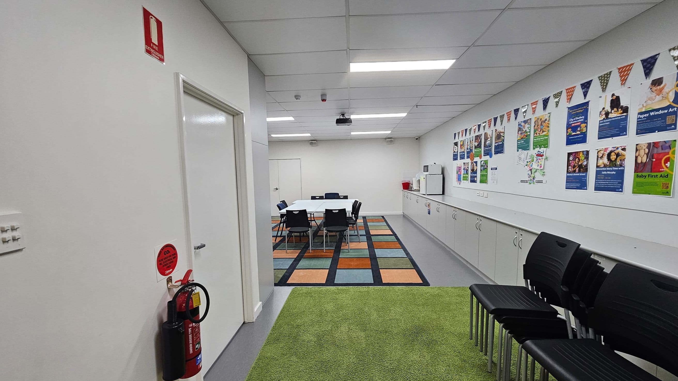 A room with a green carpet in the foreground and a colourful patterned rug in the middle. On the left, a white door with a fire extinguisher mounted on the wall. In the centre, a table surrounded by black chairs. The right wall features cabinets and a series of posters and bunting. A microwave and a red bin are on the counter. The ceiling has fluorescent lights and a projector. A stack of black chairs is on the right.