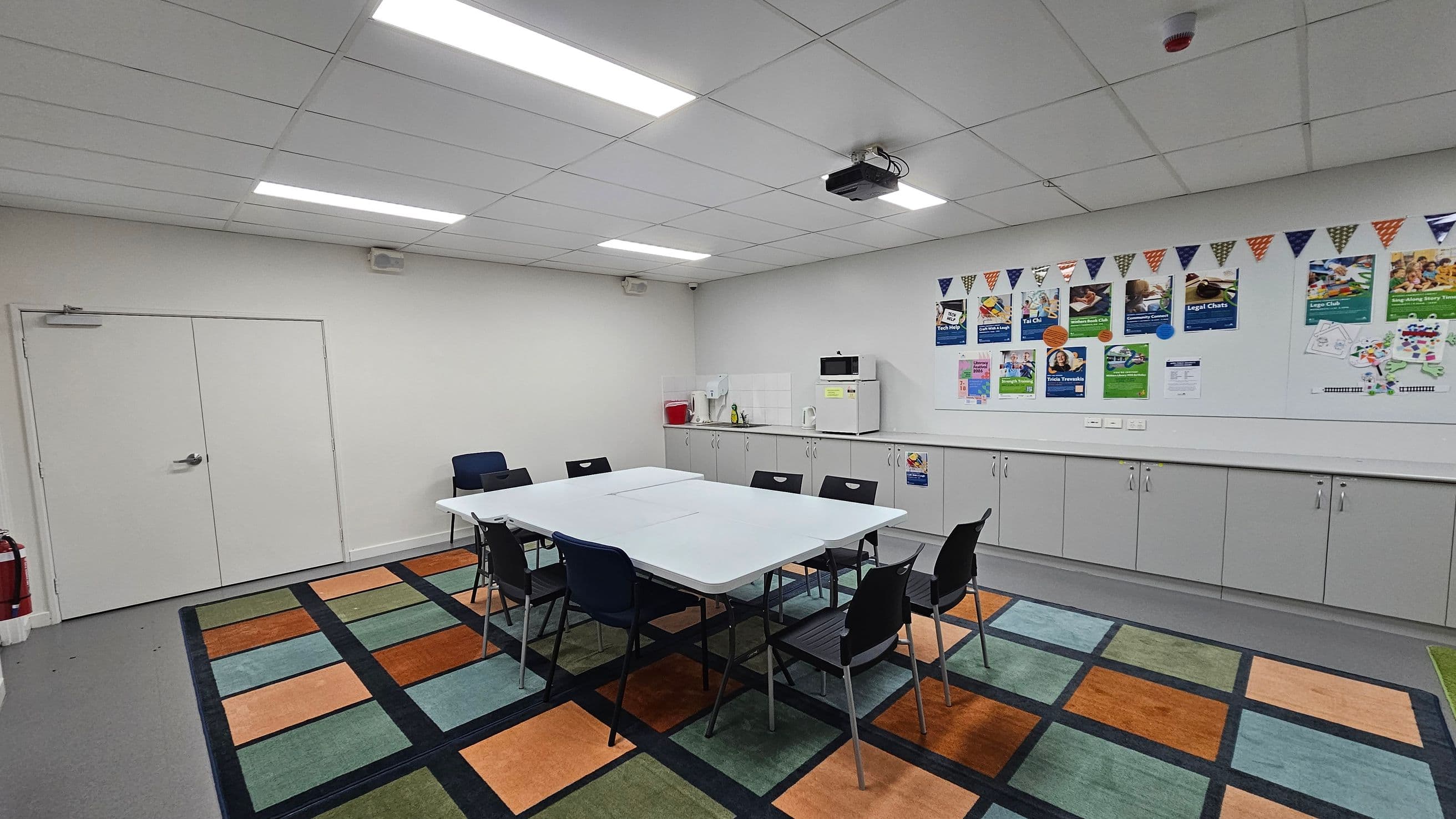 A room with a rectangular table in the centre surrounded by black chairs. The floor features a colourful, geometric-patterned rug. On the right, a long counter with cabinets below holds a microwave and a kettle. Above the counter, the wall is decorated with various posters and a string of triangular flags. The ceiling is fitted with fluorescent lights and a projector. A double door is on the left, and a fire extinguisher is nearby. The walls and ceiling are white.