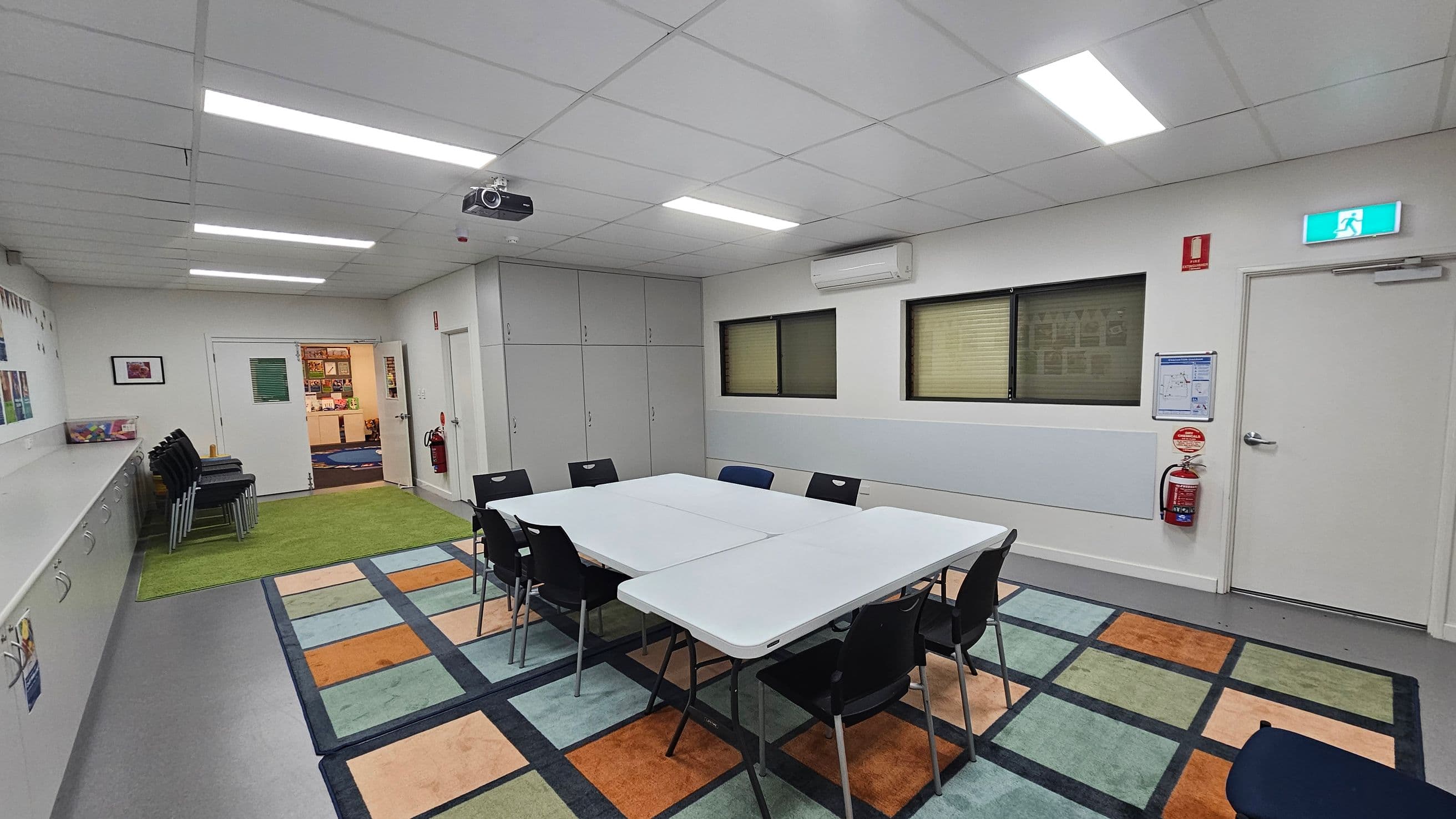A room with a white ceiling and fluorescent lights. In the centre, there are white tables arranged in a rectangle with black chairs around them, placed on a colourful, patterned carpet. On the right wall, there are two windows with blinds, an air conditioning unit, and a fire extinguisher near an exit door with a lit sign. The left side has a green carpet, stacked chairs, and a counter with cabinets. The back wall has a doorway leading to another room.