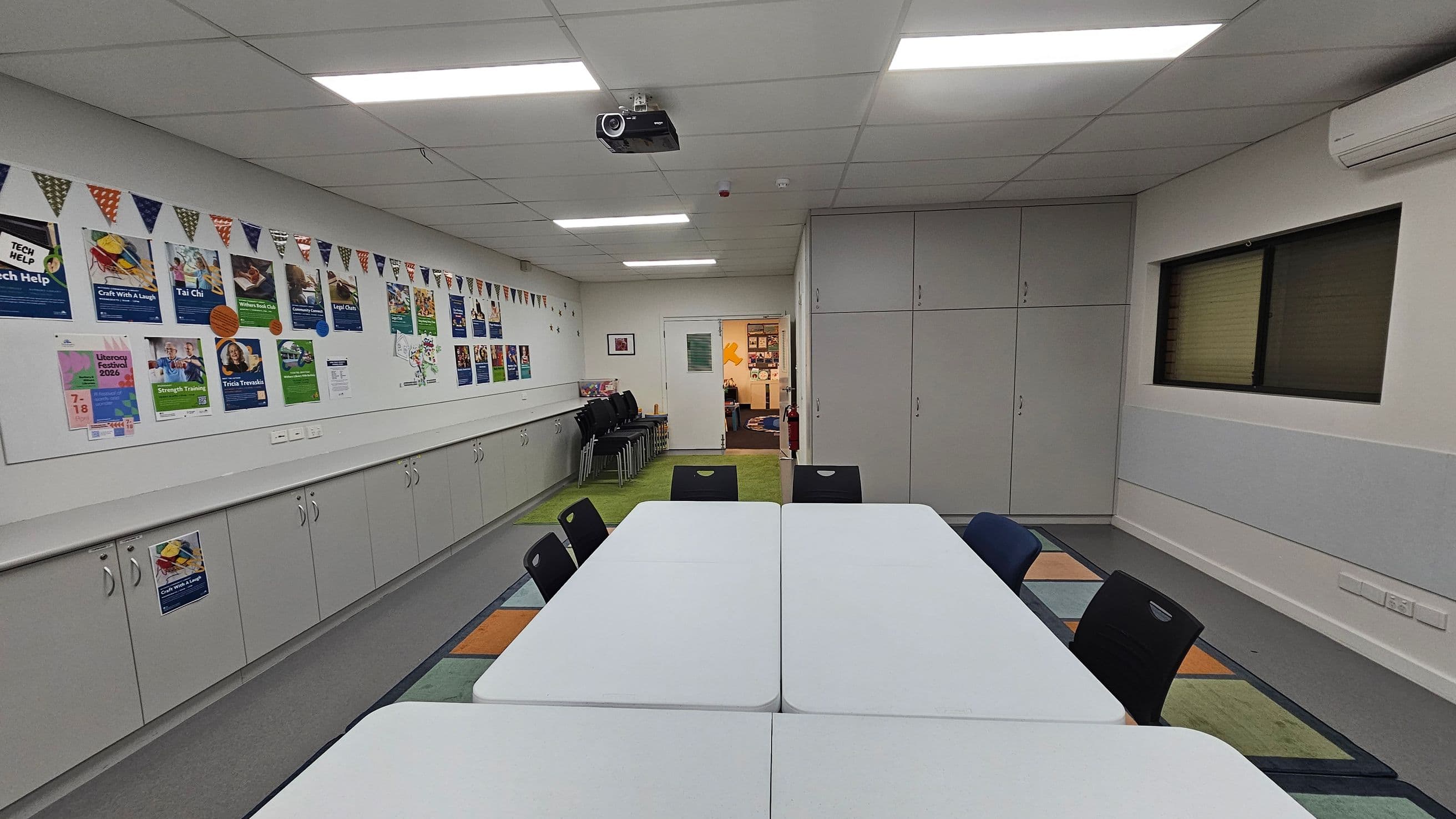 A room with a white ceiling and fluorescent lights. On the left, a long wall features colourful posters and a string of triangular flags above grey cabinets. In the centre, white tables are arranged in a rectangle with black chairs around them. The right side has a window with closed blinds and more cabinets. The floor is covered with a multicoloured carpet. At the back, an open door leads to another room with visible decorations. A projector is mounted on the ceiling.