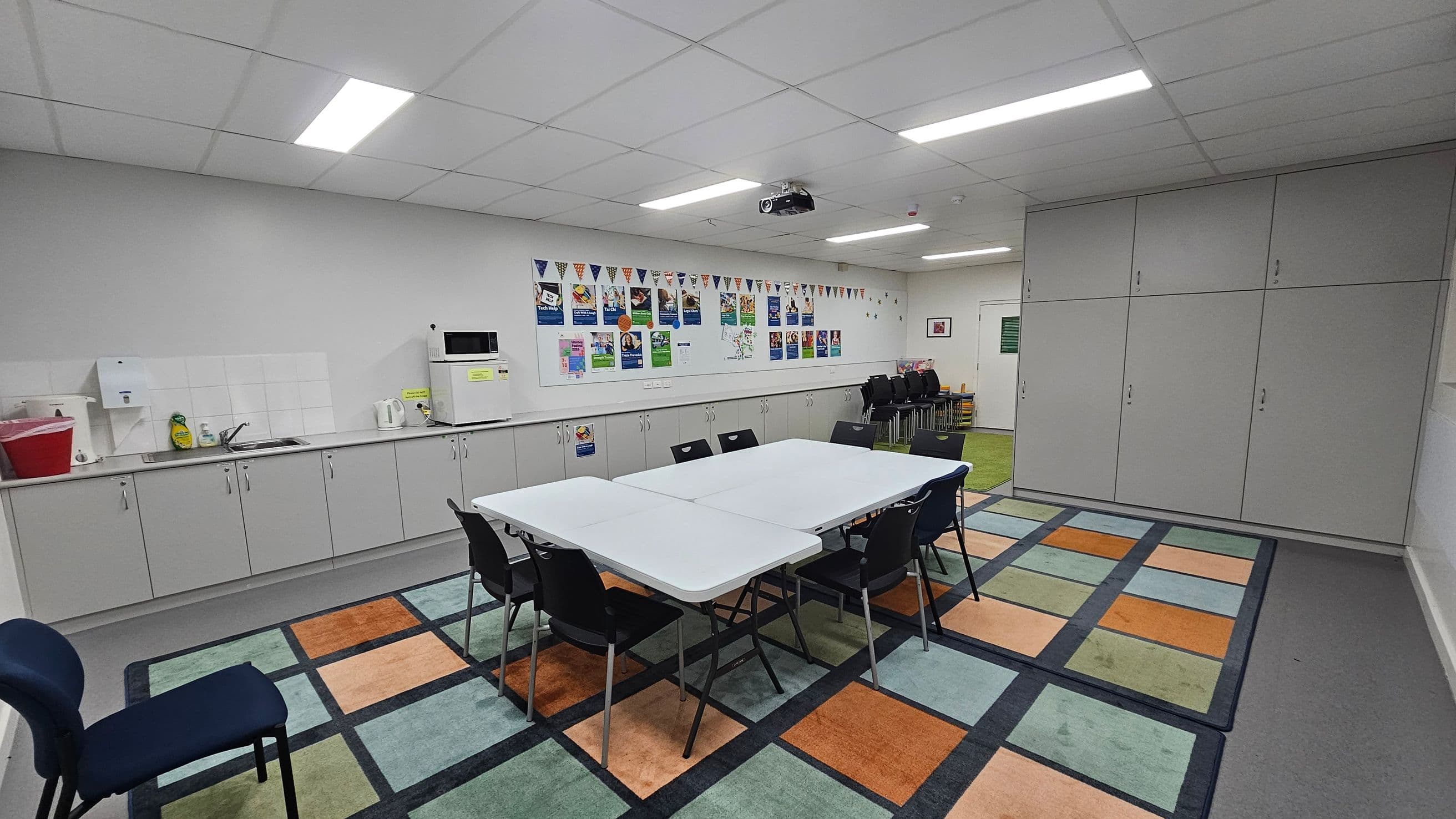 A room with a colourful checkered carpet featuring orange, blue, and green squares. In the centre, there are white tables arranged in a rectangle with black chairs around them. On the left, a kitchenette with grey cabinets, a sink, a microwave, and a kettle. The back wall has posters and bunting. On the right, large grey storage cabinets. The ceiling has fluorescent lights and a projector.