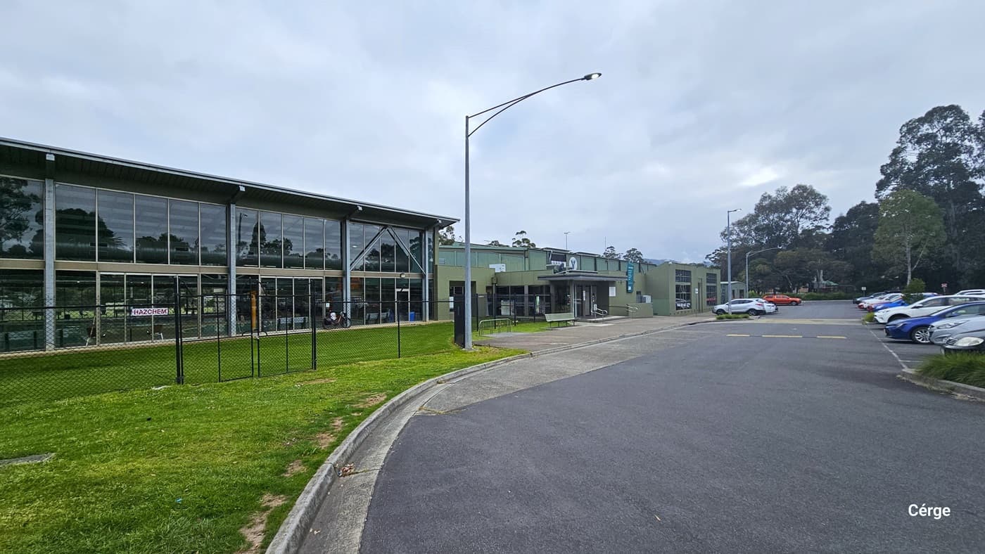 his parking area, a short walk from Yarra Centre, accommodates cars of various models and colors in designated slots, with some in motion. A tall street light stands on the right, near a bush. The view provides a comprehensive view of Yarra Centre, a substantial commercial building constructed with brick, glass, and steel. Tall trees and green grass surround the area