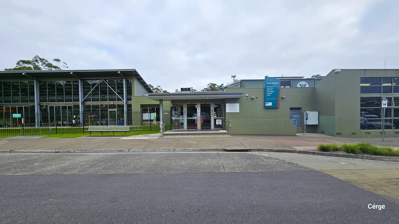 A closer view of the entrance of Yarra Centre. The building, constructed with concrete, glass, and steel, features a green "Yarra Centre" signage on the right wall. A short step is present at the entrance, and to the right, there's a ramp for accessibility, marked with signage. Additionally, a concrete plant box with ornamental plants is on the right, and on the left, a bench is provided for seating.