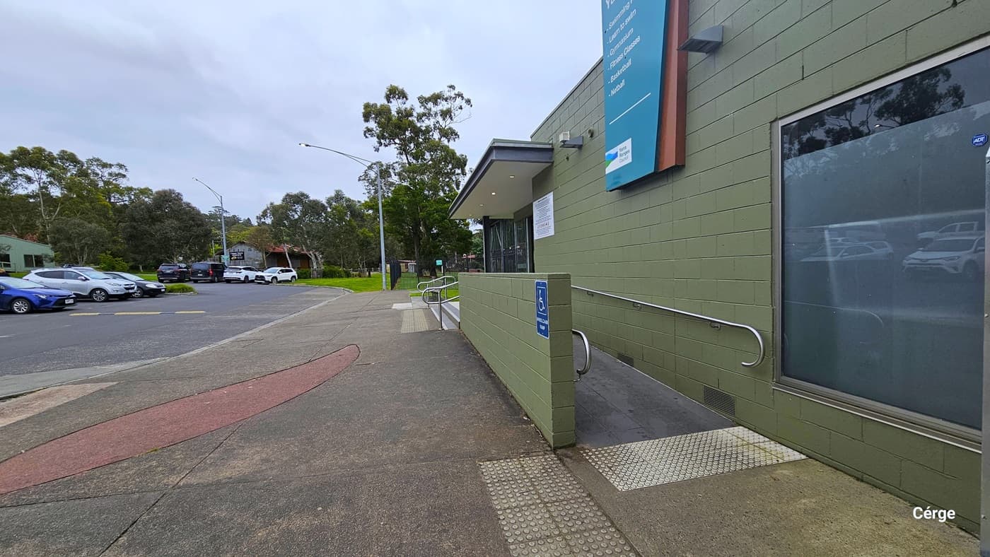 The flooring in this area is slightly uneven. An accessible entrance ramp with metal railings and signage is on the right, and white truncated domes ensure safety before the ramp. To the left is an olive-colored building with a large glass window, featuring a "Narra Centre" sign on top. On the left, a parking area hosts a few cars. The pathway leading to the entrance is designed in an oval pattern.