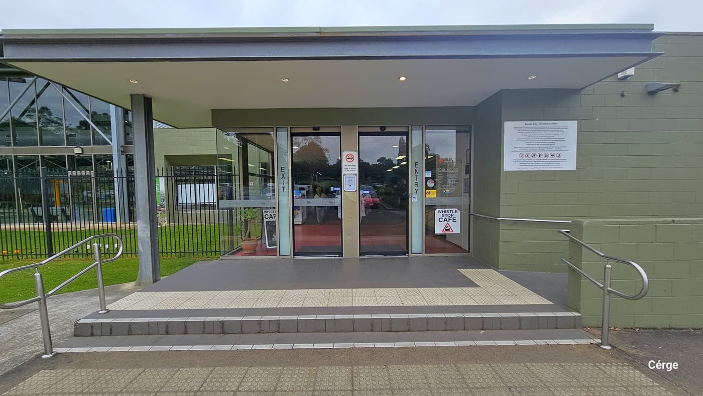 The entrance to Yarra Centre has a main entrance with two steps, featuring truncated domes below for safety. Metal railings are present on both ends. In the middle, a glass door is marked with "Entry" on the right and "Exit" on the left. A banner on the green wall displays reminders and safety instructions. Additional instructions and banners are affixed to the glass door and window. To the left, a section of the building is visible, surrounded by a fence, and adjacent to a grassy field.