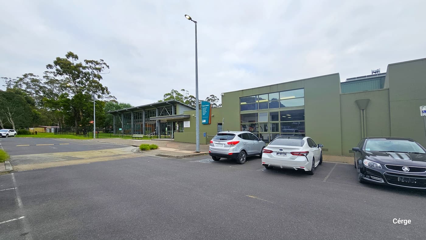 This area displays accessible parking, a short distance from the entrance. Clear signage marks the designated accessible parking spaces. To the right is the front of the building, featuring concrete walls painted in olive green and glass windows. Strategically placed tall streetlights illuminate the area. Concrete plant boxes with green ornamental plants enhance the surroundings. Trees and a white car are also visible in the distance.