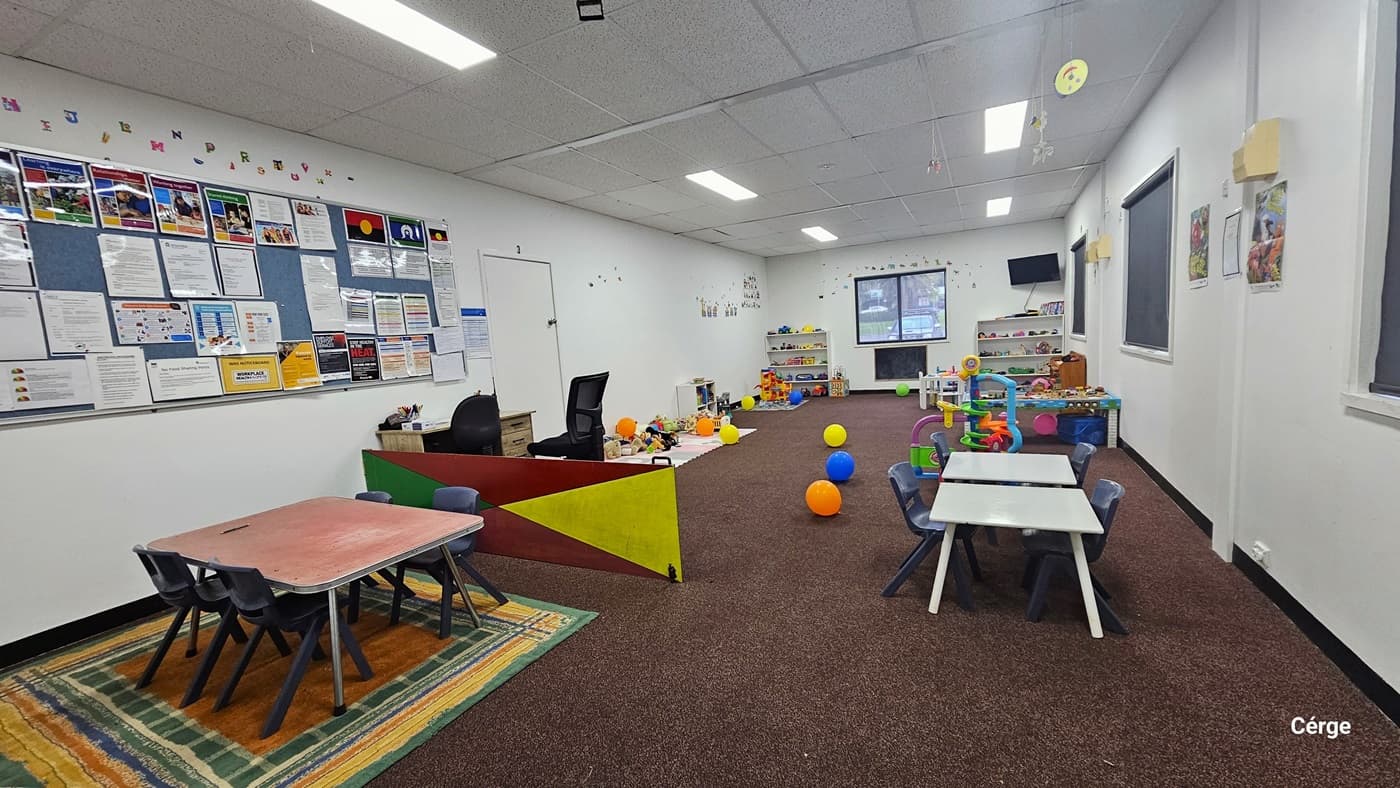 An open door marked "Multi-Purpose Room 1" provides access to the Creche. A sticker and a poster are affixed to the glass door. To the right is a registration desk. A low gate secures the area, with a safety reminder posted on it. Inside, the floor is carpeted with a rubber mat, and tables, chairs, and children's toys are visible. The walls are painted white, including the ceiling which has a porous design. A green exit sign hangs on the ceiling.