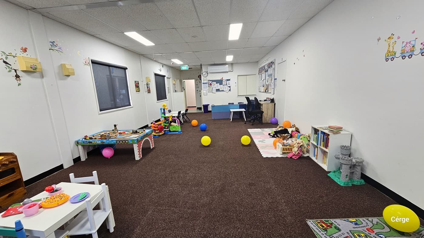 Inside the Creche, an office desk with chairs is on the right, adjacent to a kids' table with chairs placed on a floor mat. The left wall is adorned with posters containing reminders, educational materials, and colorful imagery. Two windows are situated on the left side. Tables and chairs with toys atop are lined up near the left wall, and the floor is covered with a brown rubber mat. To the right, playing mats filled with toys are present, along with a display cabinet housing books.