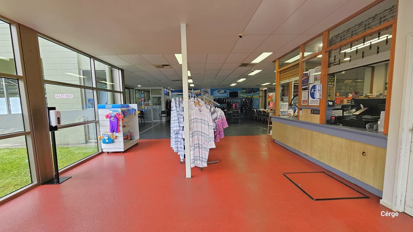 Upon entering the Yarra Centre, the reception area is to the right. The reception features a wooden design with grey lining on the edges, and the window is adorned with multiple banners. In the middle, a display rack showcases clothes, while on the left, another rack displays baby clothes. he floor is tiled with light orange tiles. Further inside, black chairs and tables are arranged for the cafe, and banners are stuck to the walls.