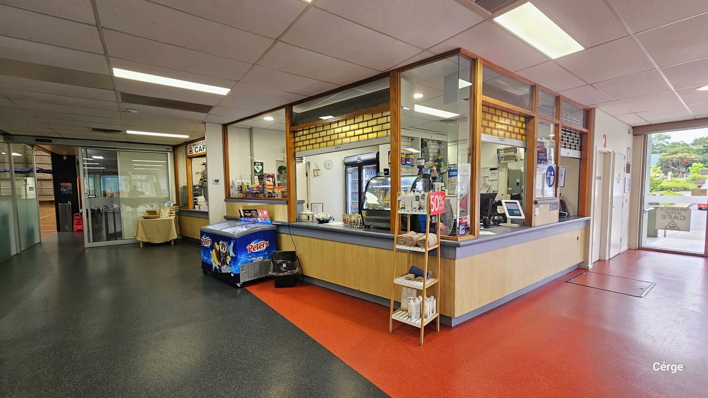 The reception desk, grey in color, features a combination of brick and lines on the front. On the desk, three computers are accompanied by swipe machines and scanners. Banners with reminders are affixed to the window. In one corner, a display rack showcases bathroom essentials with a discount rate sign. Further left, there is a retail shop and a cafe. An ice cream compartment is positioned in front, with a black rubbish bin nearby. Continuing left, a glass door is visible.
