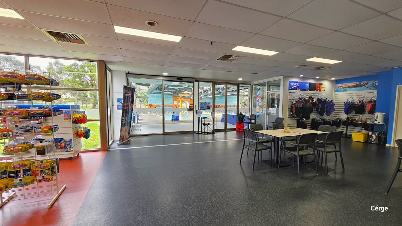 On the left stands a display rack showcasing swimming gear, goggles, and accessories. To the right is another display rack with swimwear. White tables and black chairs are placed near the cafe. A huge banner is also positioned near the glass sliding door. More banners are displayed on the racks. A mannequin wearing shorts is also visible. This area has black floor tiles on the right and light orange tiles on the left.