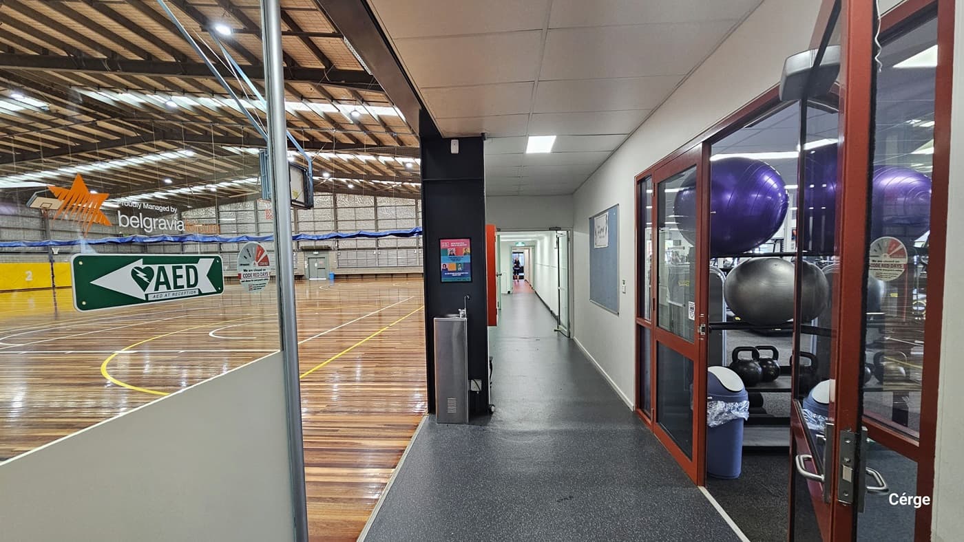 On the left of the lobby is the Sports Stadium. The floor is made of timber with yellow and white markings for various ball games. Sticker logos adorn the glass wall. To the right is the gym with training equipment, and a rubbish bin is placed near the door. Along the corridor is a drinking fountain, and signage is displayed along the corridor.