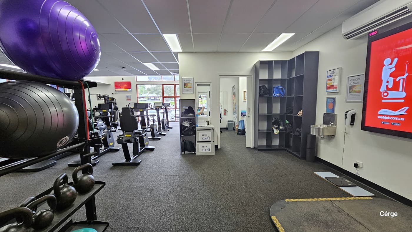 Inside the gym, upon entry, a display rack showcases purple and black stability balls on the left, with neatly lined-up kettlebells underneath. To the right, there's a storage area for bags, a washbasin, and an alcohol dispenser affixed to the wall. Large safety posters, along with others, are displayed on the wall. Further in, cardio machines are lined up, and a flat-screen is mounted on the left wall. The ceiling is white, and the floor is made of black rubber tiles.