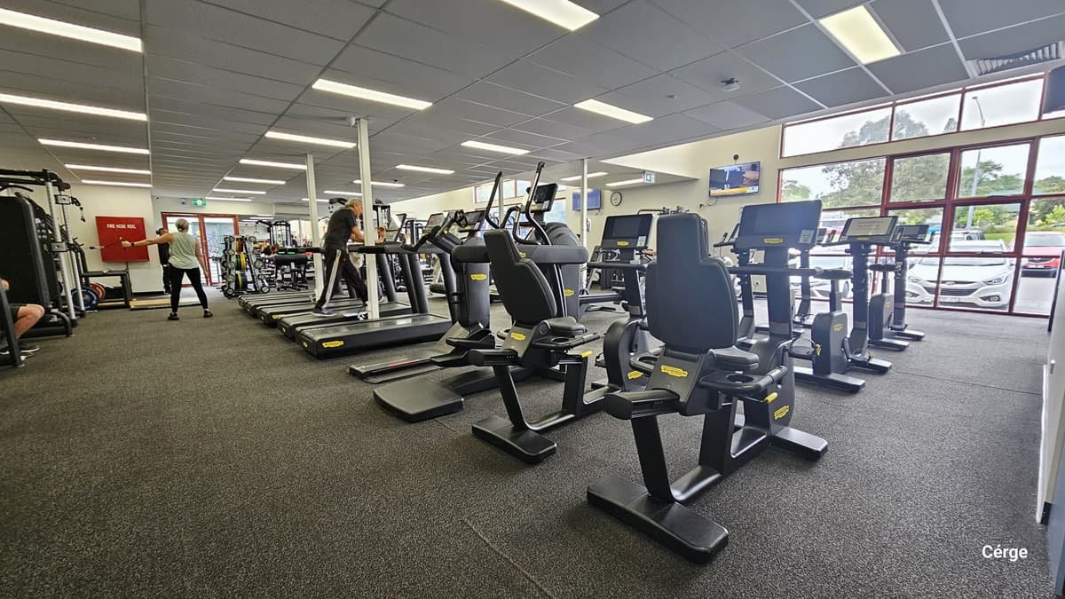 Inside the gym, various cardio machines, including treadmills, stationary bikes, and elliptical machines, are lined up in the middle. More machines are situated at the far end. The floor is covered with black rubber tiles, and the ceiling is white. Flat-screen TVs, including a round wall clock, are mounted on the walls. On the left, a fire hose reel is affixed to the wall. Some individuals are seen in motion. On the right, the glass window provides a view of the car park outside.