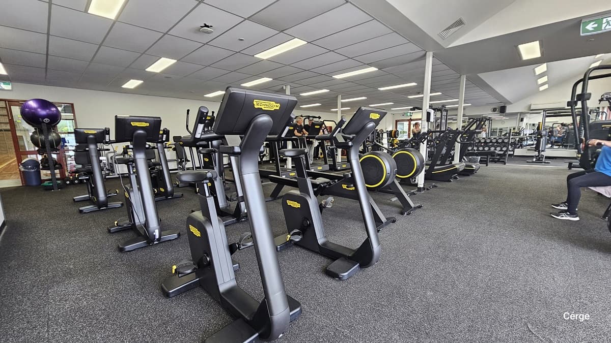 Inside the gym, a variety of cardio machines, such as treadmills, stationary bikes, and elliptical machines, are arranged in the middle, with additional machines at the far end. The floor is covered in black rubber tiles, and the ceiling is white. On the left is the door entrance, where stability balls, kettlebells, and a rubbish bin are visible. Some individuals are seen in motion.