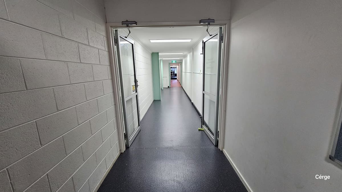 This is the Cardio Room, a spacious area with floor-to-ceiling mirrors adorned with LED lights on the left. The instructor stage is positioned behind, near the glass window. A storage with cleaning accessories is visible. On the wall, there's a round mirror above the glass window, accompanied by a flat-screen TV and a speaker. The floor features black rubber mats, and the ceiling is white with a porous design.