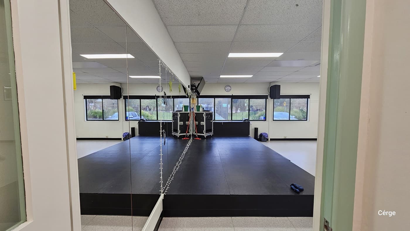 In the cardio room, a spacious area features a black rubber mat on one side and a rack with neatly arranged weight equipment on the right. A wall fan is placed nearby. In the middle, a storage area neatly displays various accessories like medicine balls, gloves, and resistance ropes. A glass window and framed decorations are on the left wall. The space is predominantly white, with a porous ceiling design and a centralized air conditioning system overhead.