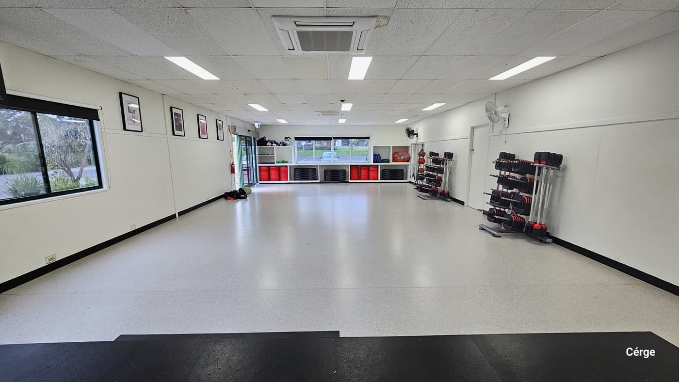 In the cardio room, a spacious open area features a centrally located training stage. On the left, a neatly arranged rack displays various weight equipment, accompanied by a wall-mounted flatscreen TV and speakers. Towards the far end, various accessories are showcased. On the right, a glass window with a round clock affixed to it allows natural light. The entire area is white, including the ceiling with a porous design.