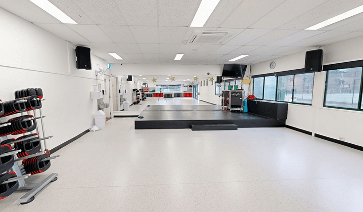 In the cardio room, a spacious open area features a centrally located training stage. On the left, a neatly arranged rack displays various weight equipment, accompanied by a wall-mounted flatscreen TV and speakers. Towards the far end, various accessories are showcased. On the right, a glass window with a round clock affixed to it allows natural light. The entire area is white, including the ceiling with a porous design.