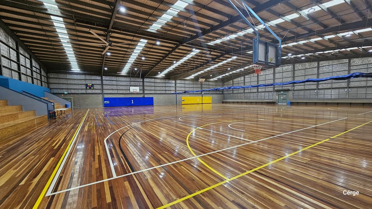 Yarra Sports Stadium boasts timber floors marked with colourful indicators for various ball games. The pitched ceiling is adorned with fluorescent lighting and large fans. Tiered seating is on the left, and distinct blue and yellow buntings mark the two courts at the far end. A drinking fountain is located near the glass door entrance, and posters and stickers adorn the walls.