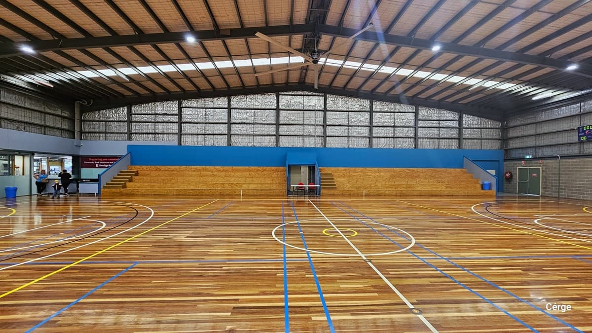 The Yarra Sports Stadium. The timber floors are marked with colourful indicators for various ball games. The pitched ceiling is adorned with fluorescent lighting and a large fan. In the center, tiered seating is positioned against the wall. On the left corner, there is an office with two desk tables and a banner. Insulation material is installed from the upper wall to near the ceiling. On the right is a green door and a digital screen affixed to the wall.
