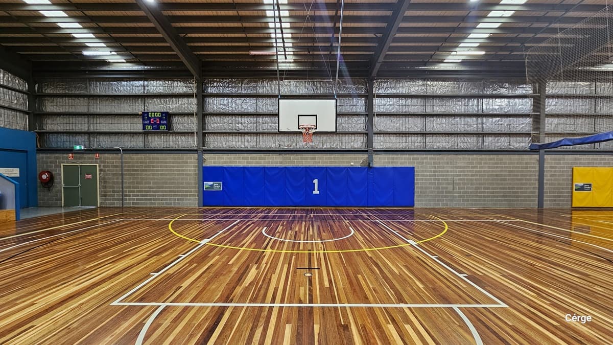 This offers another view of Yarra Sports Stadium. The timber floors feature colourful indicators for various ball games. The pitched ceiling is adorned with fluorescent lighting. Blue and yellow buntings with numbers distinguish between the two courts. In the middle, a basketball board is visible. To the left is a green door on a brick wall, and a digital screen is affixed to the wall.