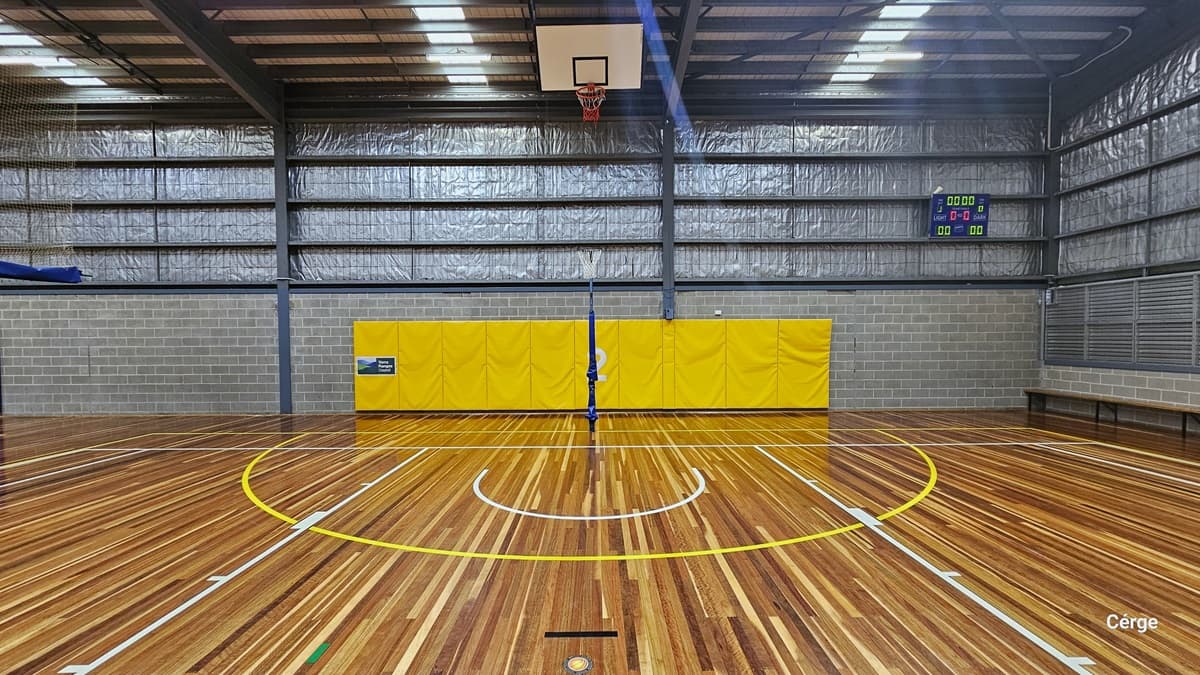The Yarra Sports Stadium. The timber floors are marked with colourful indicators for various ball games, complemented by a pitched ceiling adorned with fluorescent lighting. Blue and yellow buntings with numbers distinguish between the two courts. In the middle, a basketball board is visible. To the left, a green door is set against a brick wall, and a digital screen is affixed to the wall. Insulation foams are also installed on the wall near the ceiling.