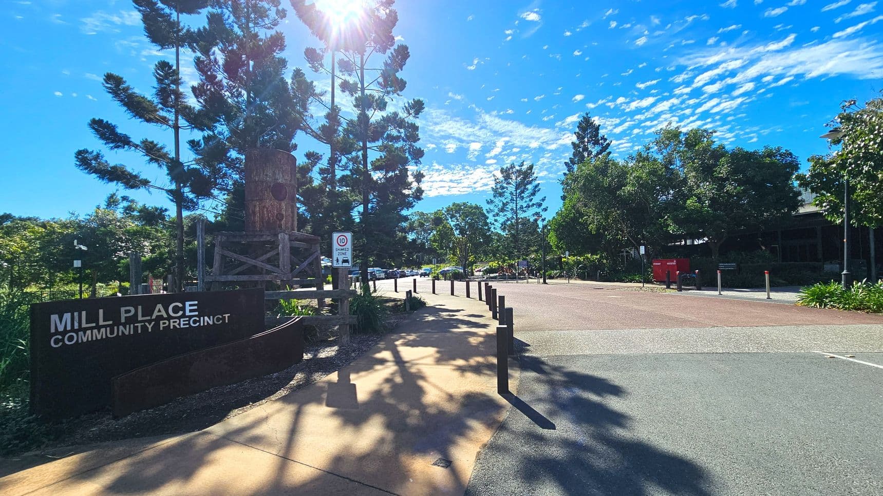 A paved pathway leads into the Mill Place Community Precinct, marked by a prominent sign on the left. Tall trees line the path, casting shadows on the ground. A wooden structure stands beside the sign. The area is open, with bollards lining the path and a speed limit sign visible. To the right, a building is partially obscured by lush greenery, and a red post box is visible near the entrance.