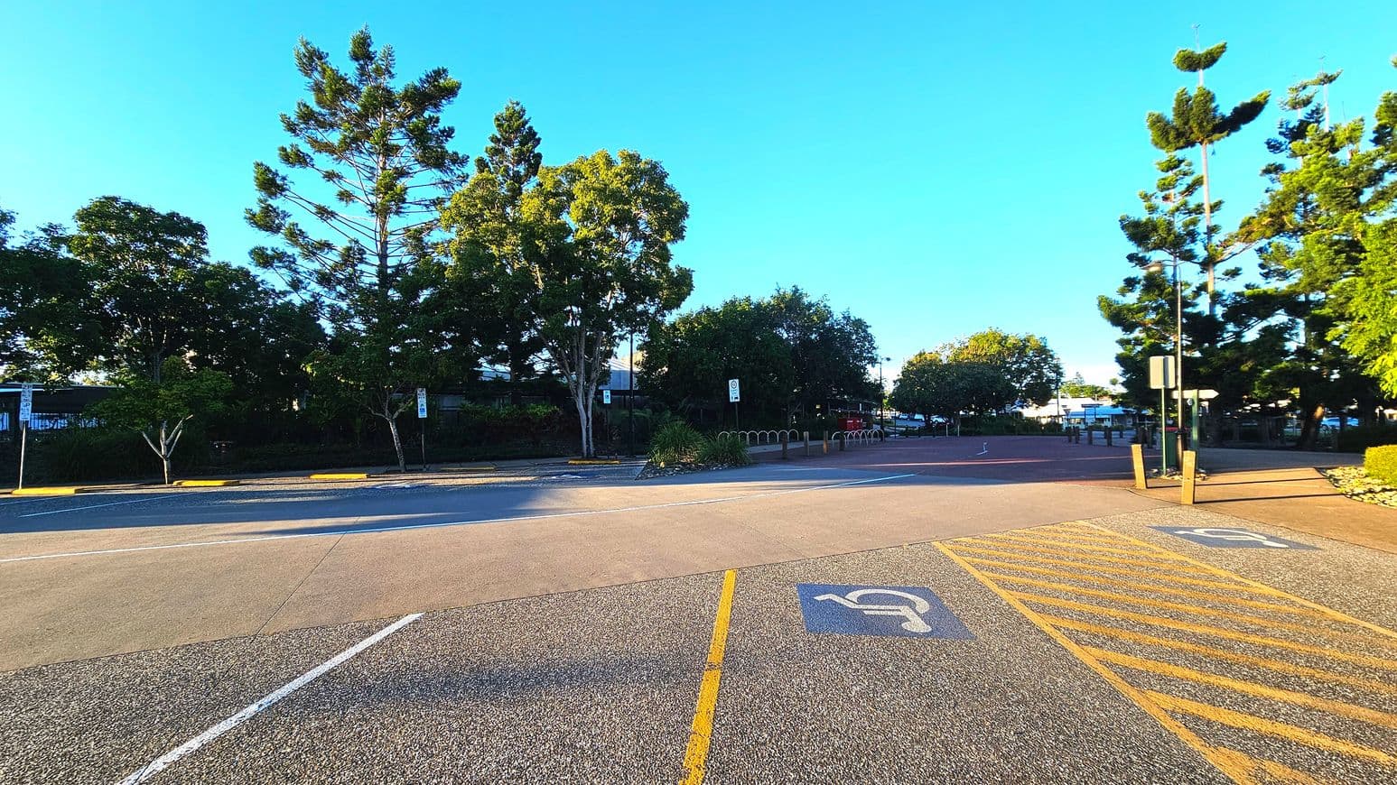A parking area with marked disabled parking spaces is visible in the foreground, featuring yellow lines and symbols on the asphalt. Beyond the parking area, a paved path leads into a tree-lined space with tall, leafy trees and a few signs. The area feels open with no visible barriers. The surface transitions from asphalt to a reddish paved path. There are bollards and a bike rack near the path, indicating pedestrian and cyclist access.
