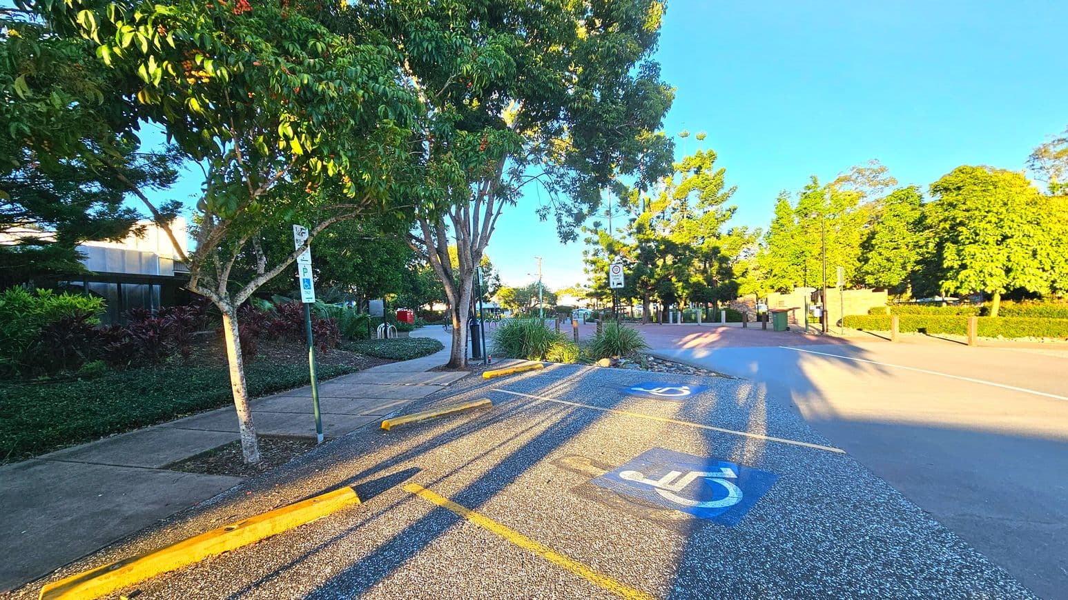 A parking area with designated disabled parking spaces marked by blue paint and white wheelchair symbols on the ground. Yellow wheel stops are positioned at the end of each space. A concrete footpath runs alongside, bordered by lush green trees and shrubs. A signpost indicates parking regulations. The area is open, with a road visible to the right and a bicycle rack in the background.