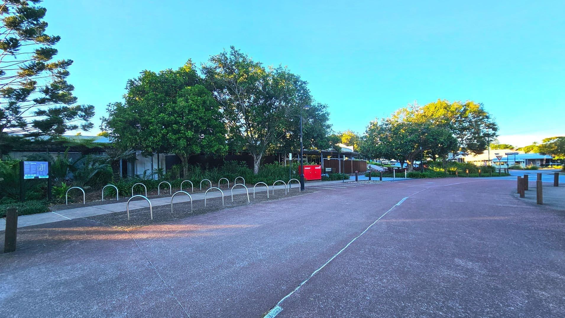 A paved pathway runs through a park-like area with metal bike racks on the left. Dense greenery, including large trees and shrubs, lines the path. A red post box and a signpost are visible near the trees. The area is open with wooden bollards marking the boundary of the path. A building with a flat roof is partially visible behind the trees, and a road with a roundabout is seen in the distance.