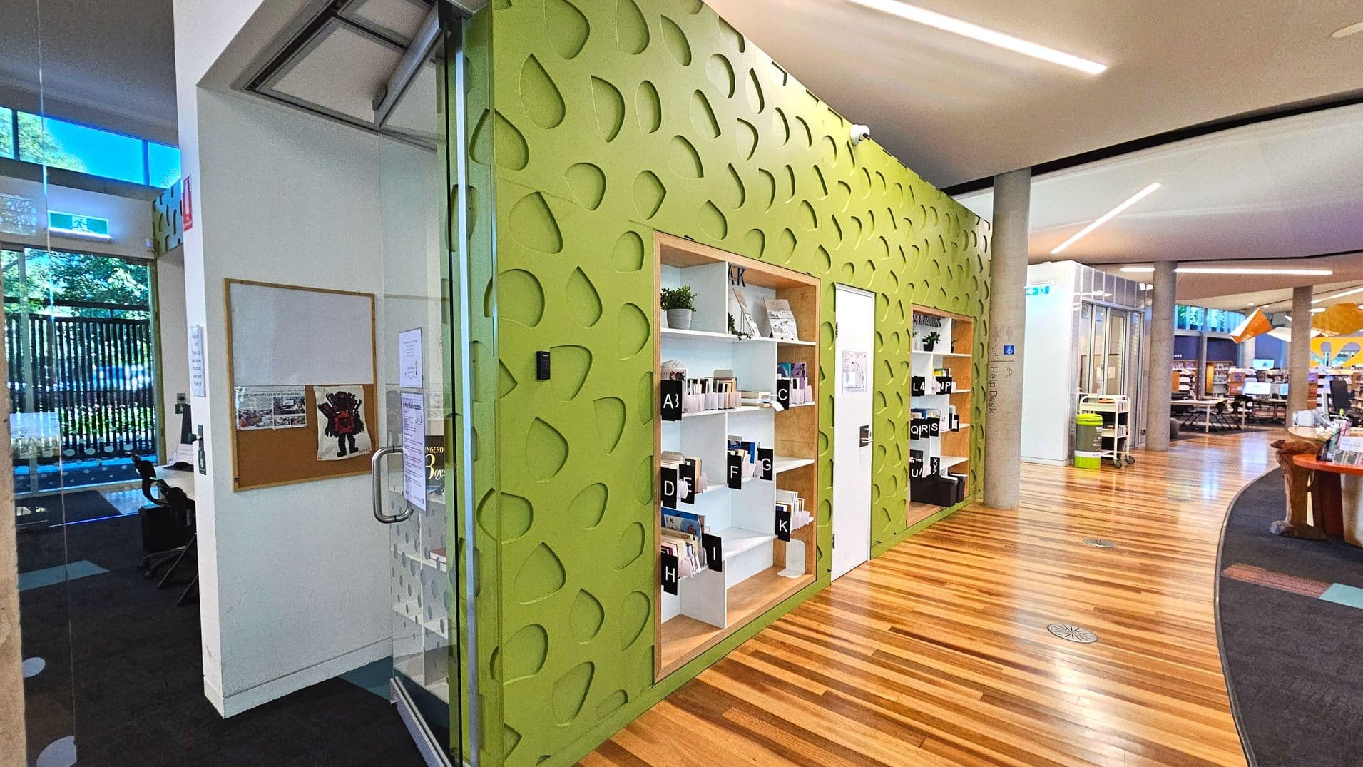 The image shows an interior space with a distinctive green wall featuring a pattern of cut-out shapes. Built-in shelves display books and small plants. The floor transitions from dark carpet to polished wooden planks. A glass door with a metal handle is visible on the left, leading to a small area with a desk and chair. The space is open with curved ceilings and columns, and a noticeboard is mounted on a white wall.
