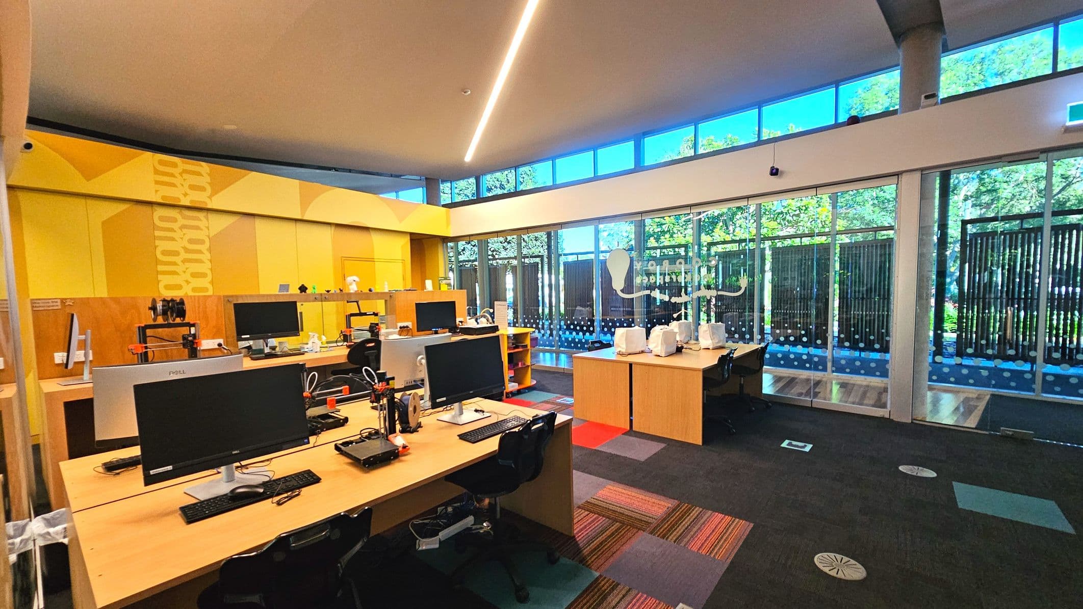 A modern workspace with several wooden desks equipped with computers and 3D printers. The room features a vibrant yellow wall with geometric patterns and a large glass wall with vertical blinds, allowing natural light to flood in. The floor is covered with a mix of patterned and solid-coloured carpet tiles. Overhead, a sleek linear light fixture runs across the ceiling, and a concrete pillar stands near the glass wall.