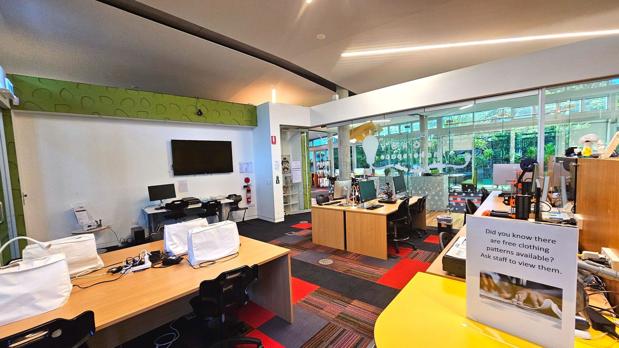 The image shows an interior space with a modern design, featuring a mix of wooden desks and black chairs. A large flat-screen TV is mounted on a white wall, with a computer station beneath it. The floor is covered with a patterned carpet in red and grey tones. A glass partition with decorative decals separates the area from a lush green outdoor view. A sign on a yellow counter mentions free clothing patterns. The ceiling is high with recessed lighting.