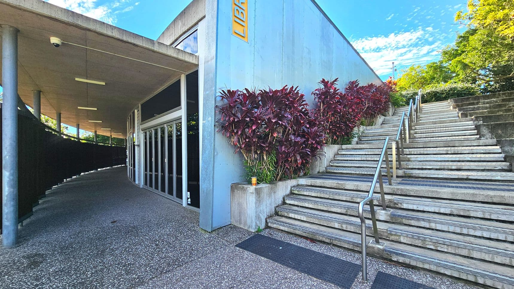 A modern building entrance with a prominent 'Library' sign on a light blue wall. To the right, a wide staircase with metal railings ascends alongside a row of red-leaved plants. The left side features a covered walkway supported by cylindrical columns, leading to glass doors. The ground is a speckled concrete surface, transitioning to steps. The area feels open with greenery visible beyond the stairs.