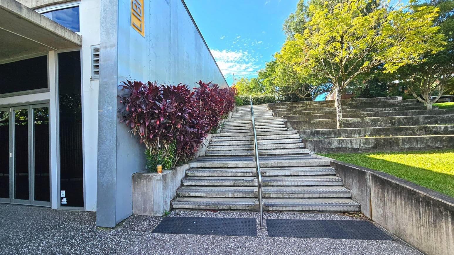 A concrete staircase with a central metal handrail ascends between a grey building on the left and a grassy area with tiered seating on the right. The building has a sign reading 'LIBRARY' and features a glass entrance. Red-leaved plants line the left side of the stairs. The ground is a mix of textured concrete and rubber mats, and a tree provides shade over the seating area.