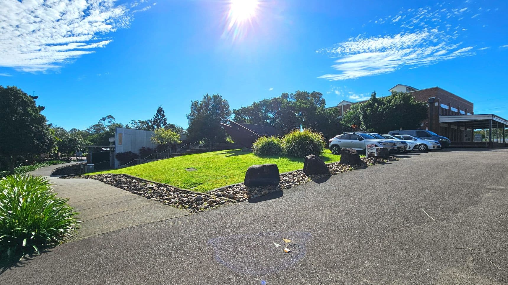 A paved area with several parked cars on the right, adjacent to a grassy slope bordered by rocks. The slope leads to a building with a brick facade partially visible in the background. A pathway curves around the grassy area, lined with shrubs and trees. Large rocks are placed at intervals along the edge of the grass. The scene is open with clear visibility of the surrounding greenery.