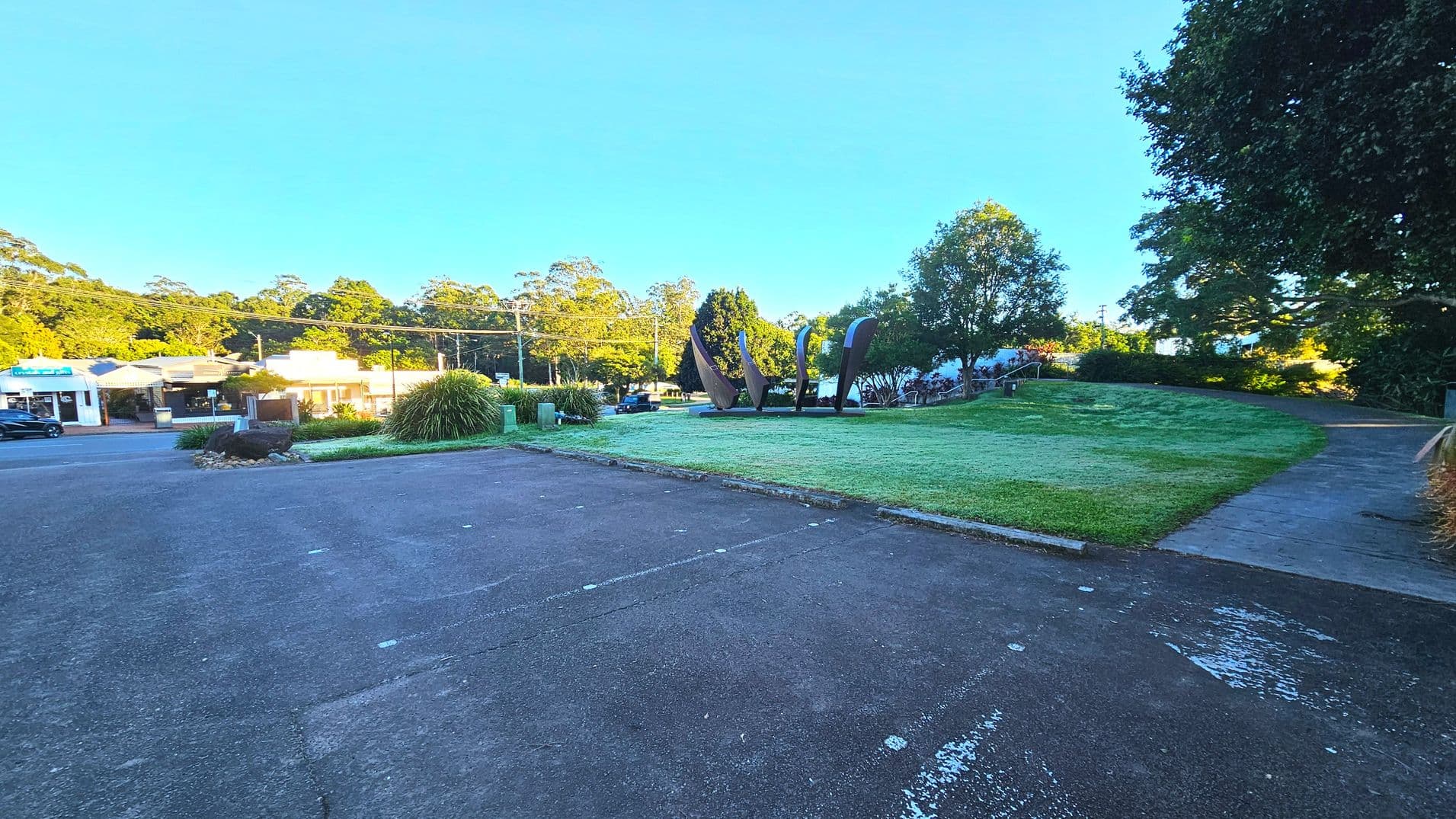 A grassy area with a curved concrete path on the right leads to a set of large, abstract metal sculptures in the centre. To the left, a paved area transitions into a road lined with small shops and parked cars. Trees and bushes surround the space, providing a natural boundary. The sculptures are the focal point, with their tall, curved forms standing prominently against the greenery.
