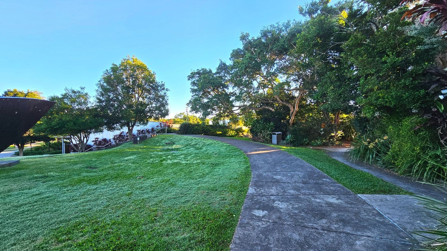 A gently curving concrete path winds through a lush garden area, bordered by well-maintained grass and dense trees. On the left, a large, dark sculpture partially appears, while a metal railing lines a small set of steps leading up a grassy incline. A bin is visible along the path, which is flanked by tall trees providing shade. The area feels open with a mix of natural and man-made elements.
