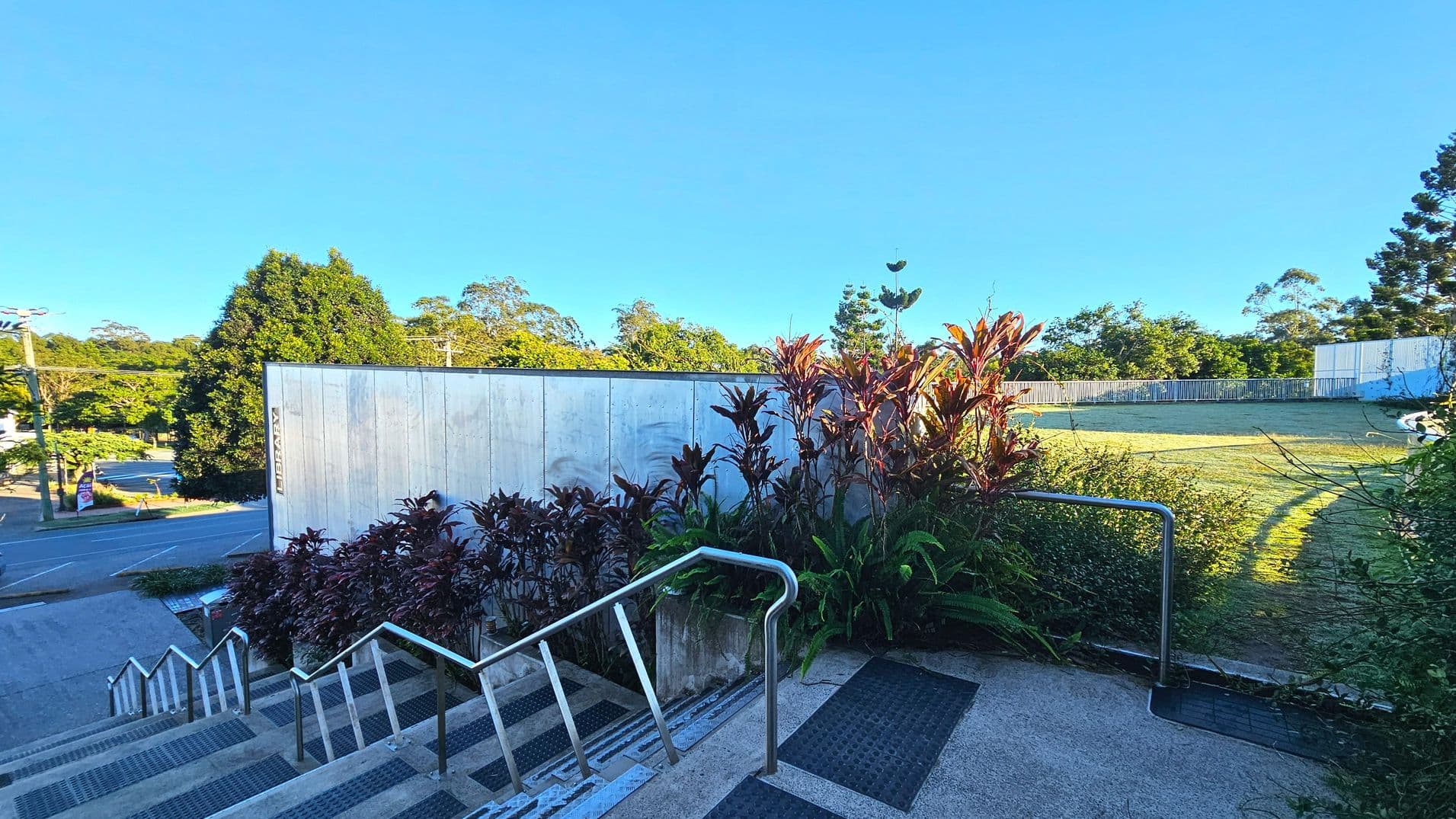 A concrete staircase with metal railings descends alongside a building with a sign reading 'Library'. The stairs are bordered by lush plants with red and green foliage. At the top, a flat concrete area transitions to a grassy lawn enclosed by a low metal fence. The scene is surrounded by trees and a road is visible to the left. The area feels open with clear pathways and natural elements.