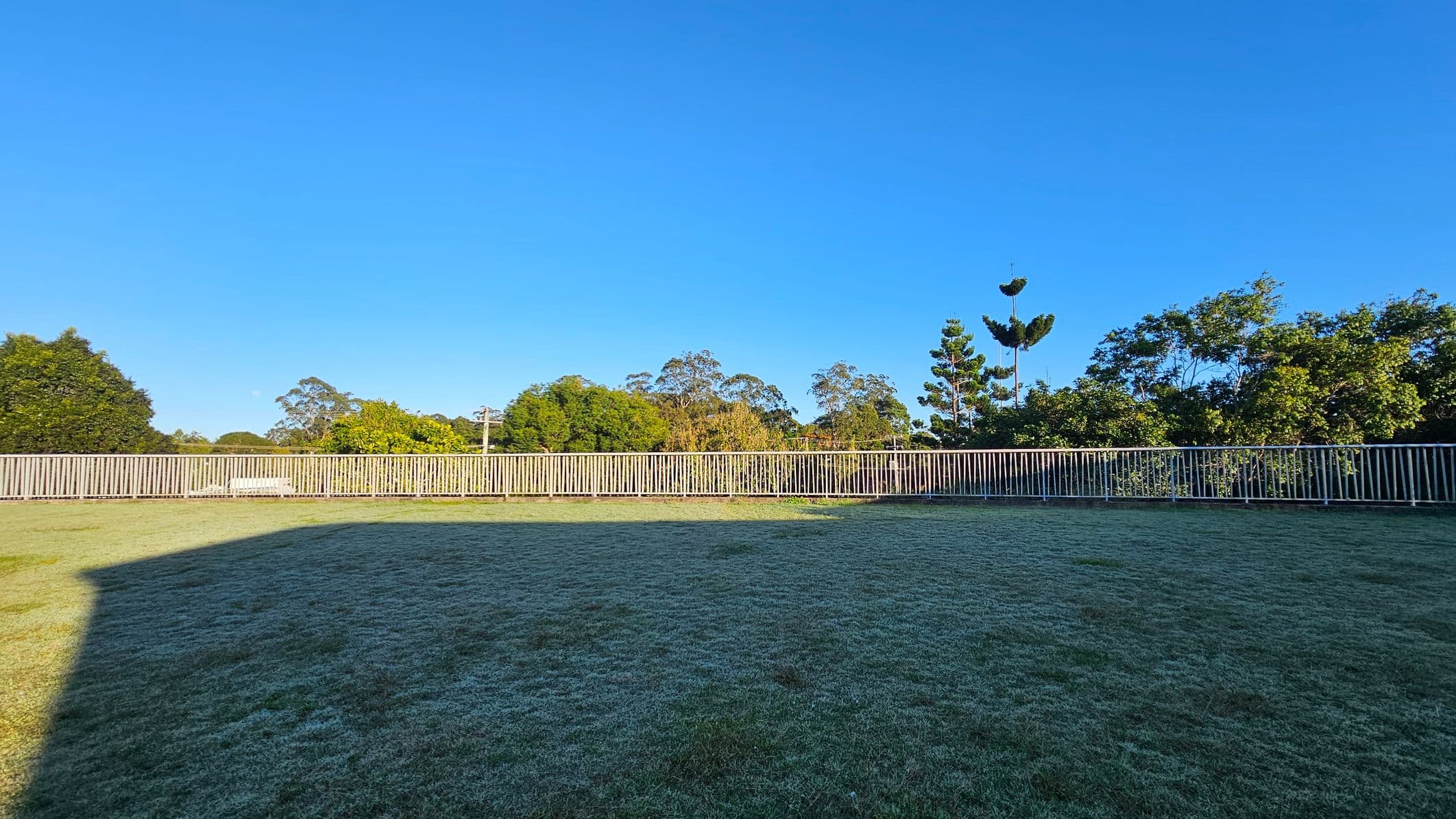 A grassy open area bordered by a long, low metal fence running horizontally across the image. Behind the fence, there are various trees and shrubs, providing a natural backdrop. The grass appears well-maintained, with a shadow cast across part of it, suggesting a nearby structure. The scene is flat and expansive, with no visible structures or pathways within the fenced area.