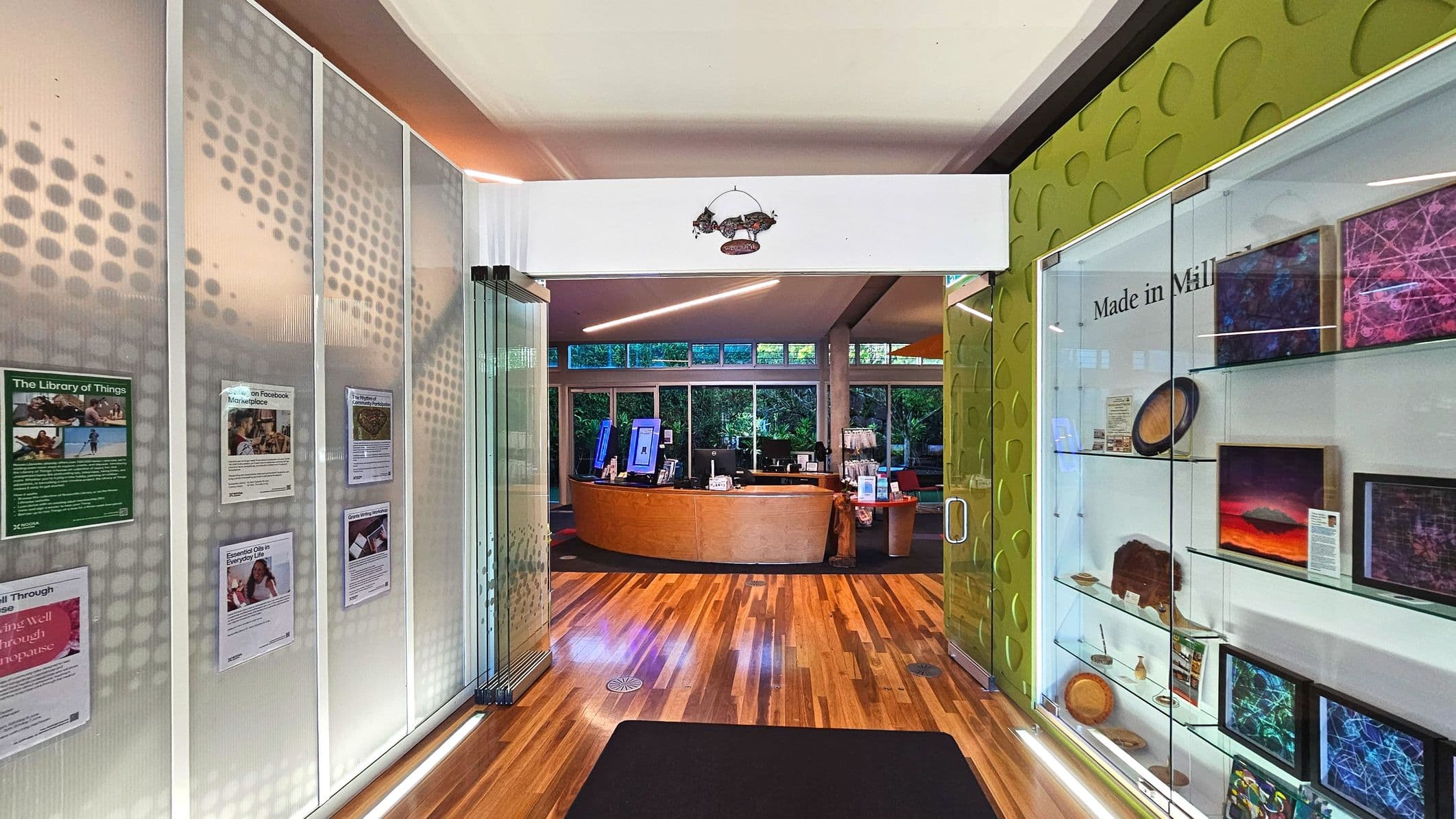 A modern interior space with a wooden floor leading to a reception area. On the left, a wall displays various posters, while the right side features a glass display case with art pieces and the text 'Made in Mill'. The reception desk is curved and wooden, with computer monitors and brochures. Large windows behind the desk reveal greenery outside. The ceiling has recessed lighting and a decorative piece hangs above the entrance.