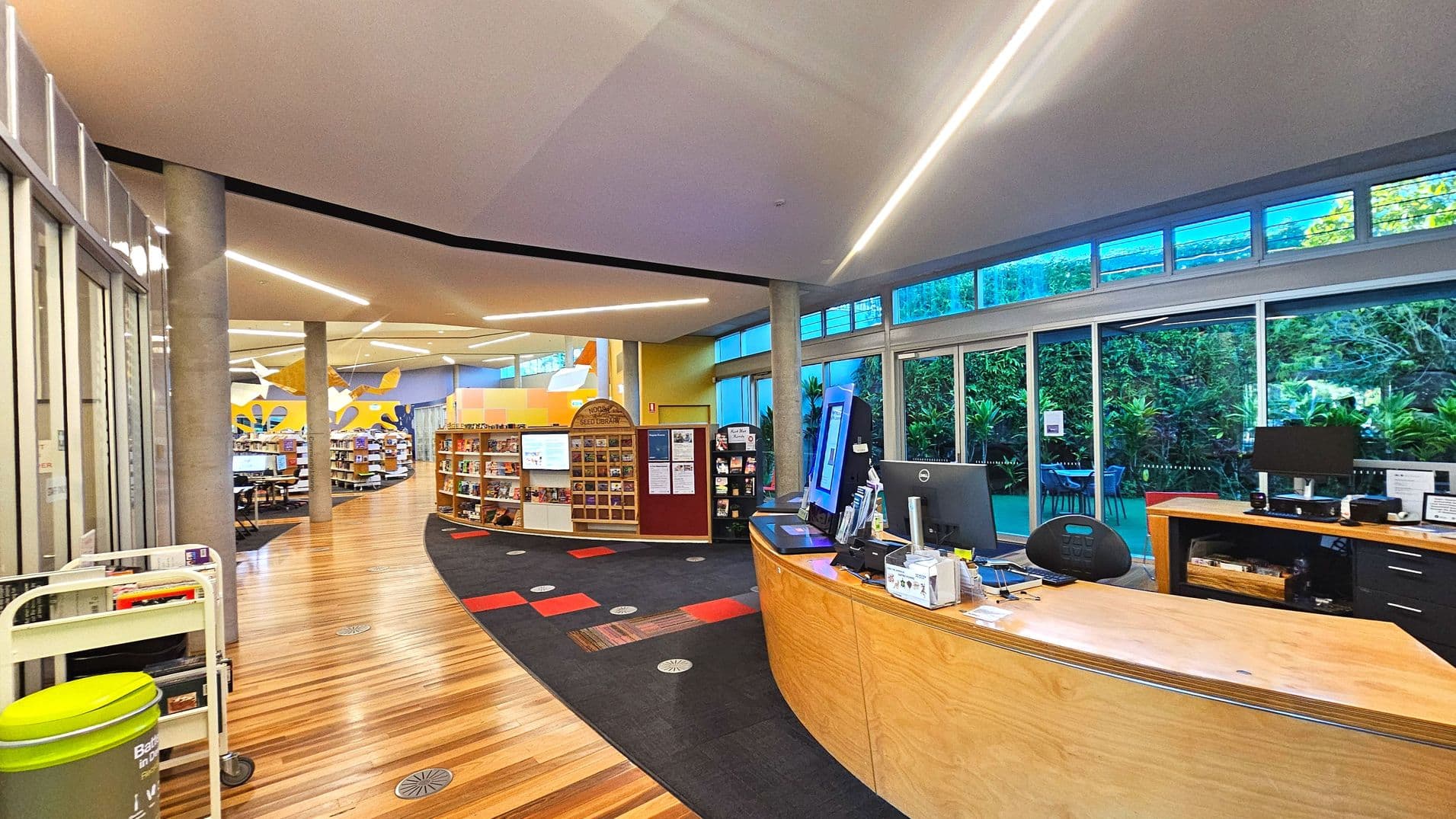 The image shows a modern library interior with a curved wooden reception desk on the right, equipped with computers and office supplies. To the left, there are bookshelves arranged in a semi-circular layout. The floor transitions from light wood to a patterned carpet. Large windows with greenery outside line the right wall, providing natural light. Overhead, sleek linear lights are embedded in the ceiling, and concrete pillars support the structure.