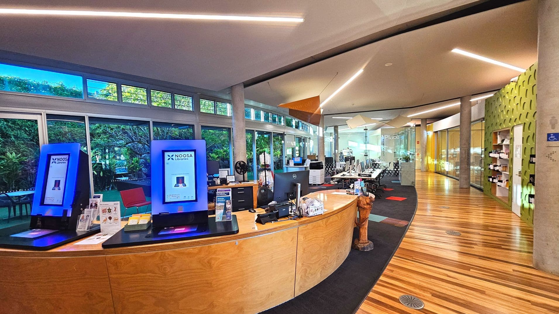 The interior of a modern library features a curved wooden reception desk with two digital kiosks displaying 'Noosa Libraries'. Behind the desk are computers and office equipment. The space is enclosed by large glass windows with greenery visible outside. The floor transitions from wood to carpet, and a green textured wall is on the right. Overhead, linear lights illuminate the ceiling, and a sign reads 'Place'.